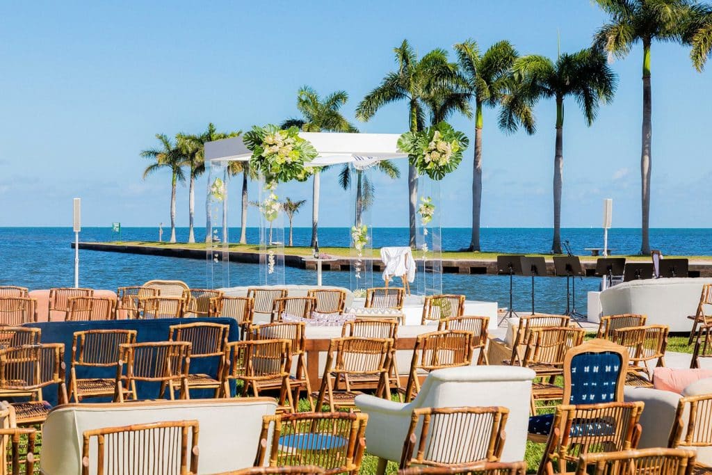 Outdoor wedding ceremony by the water with wooden chairs and palm trees
